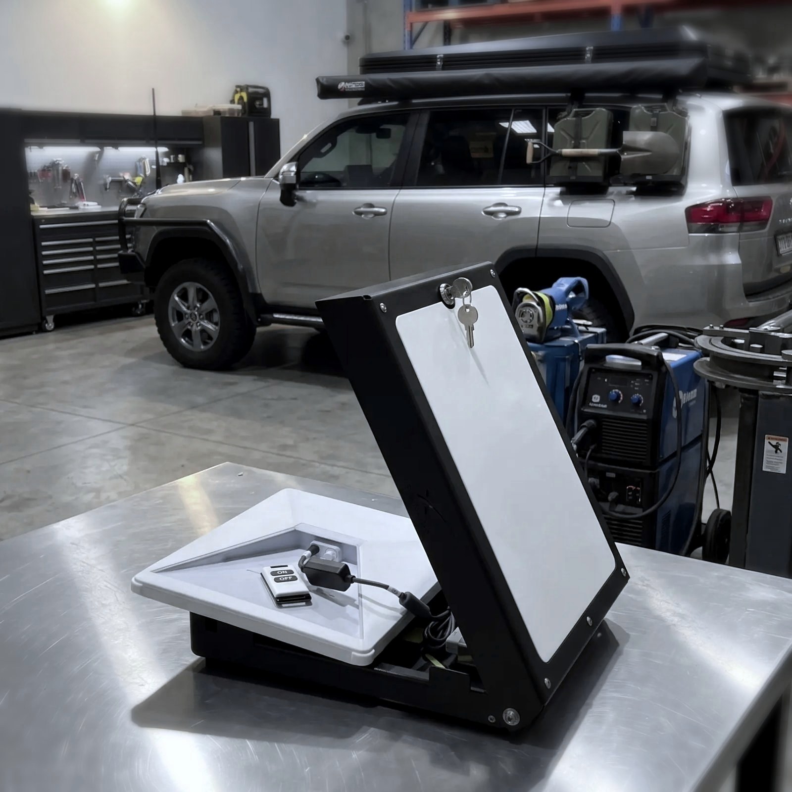 Power Hub open on a workshop bench with the lid raised and the key in the latch, the remote and inline switch wired inside, a touring LandCruiser in the background